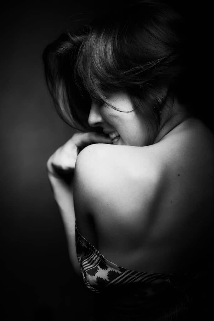 Portrait boudoir en noir et blanc d’une femme protégeant délicatement son épaule dans une lumière douce, réalisé au studio Les Délices d’un Regard à Tourcoing.